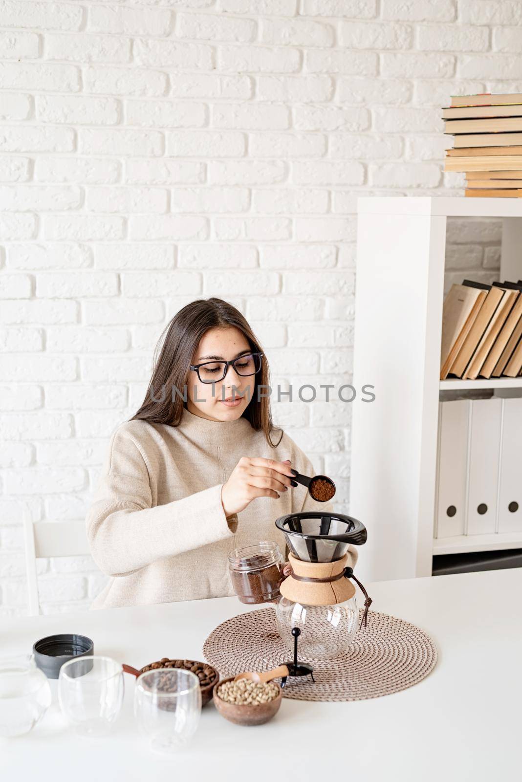 Alternative coffee brewing. Young woman brewing coffee in chemex sitting at the white table with various stuff for alternative coffee brewing, pouring grinded coffee beans into filter