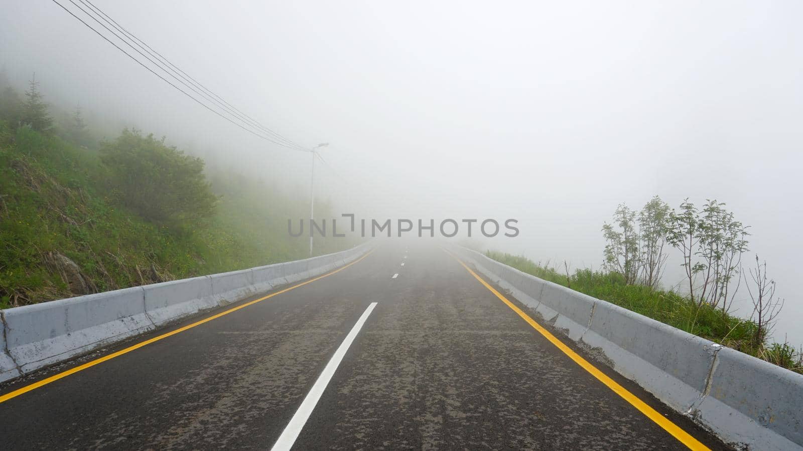 Mountain road in the fog. First-person view, walking along the road. Trees and green grass are visible along the edges. There are poles for lighting. Thick fog obscures visibility. Dividing strip.