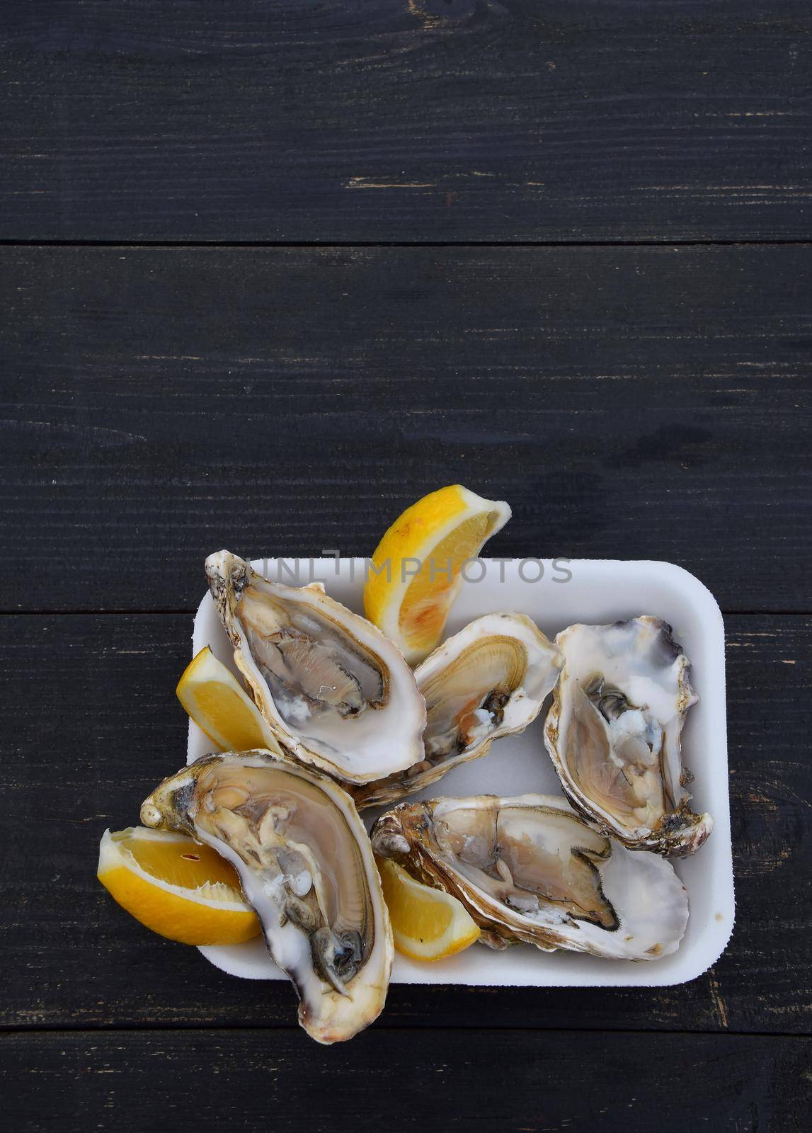 Close up portion of fresh open raw juicy oysters with lemon wedges on white plastic tray over black wooden table with copy space, high angle view
