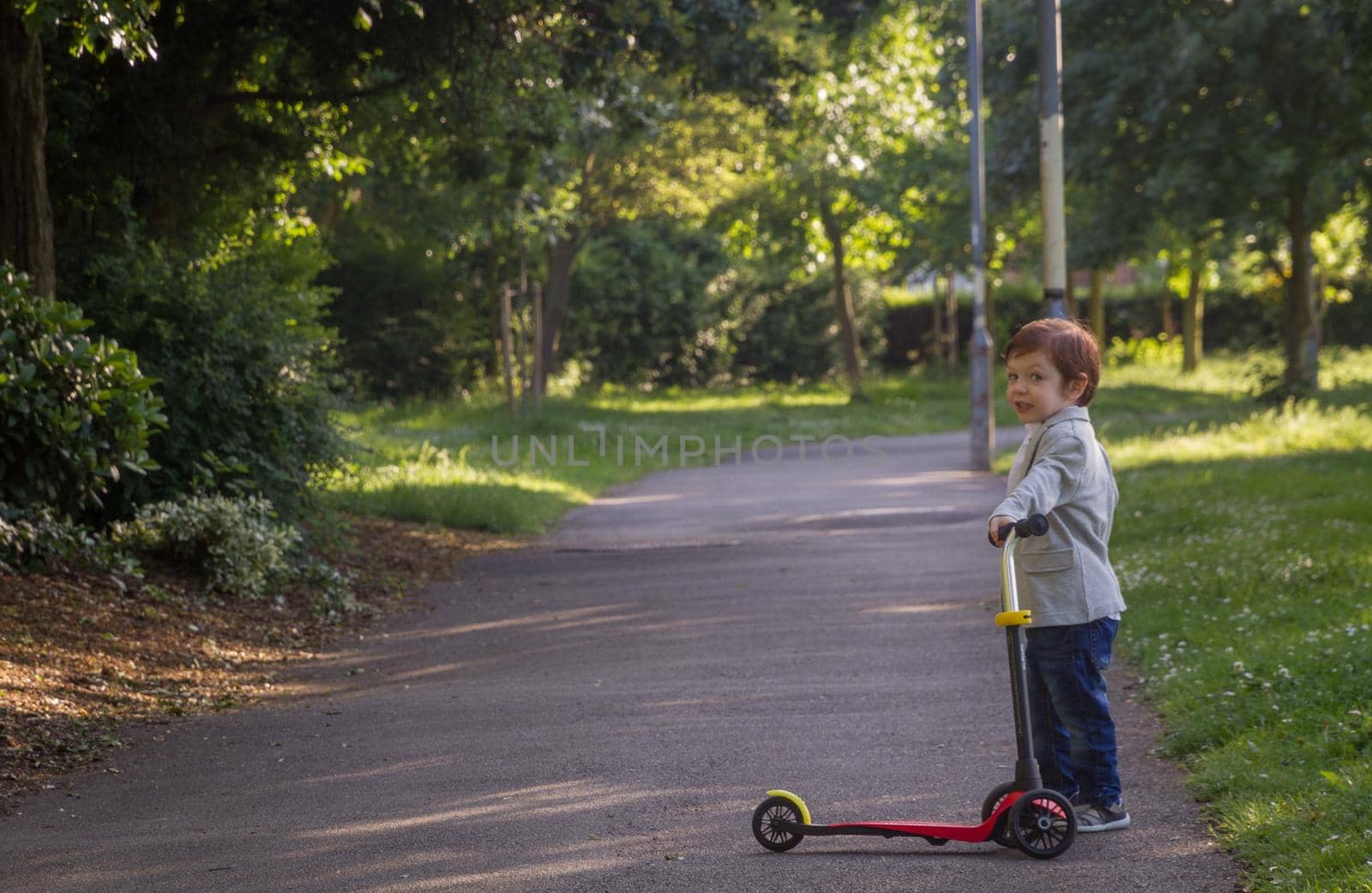 A cute, redhead, blue-eyed baby boy wearing a blazer holding a red scooter in a park on a sunny day