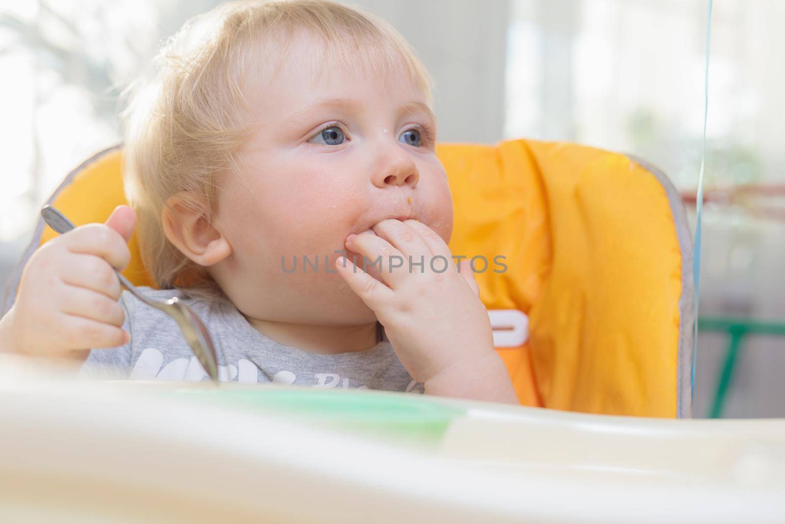 Little child sits on a high chair and eats with a spoon by Yurich32