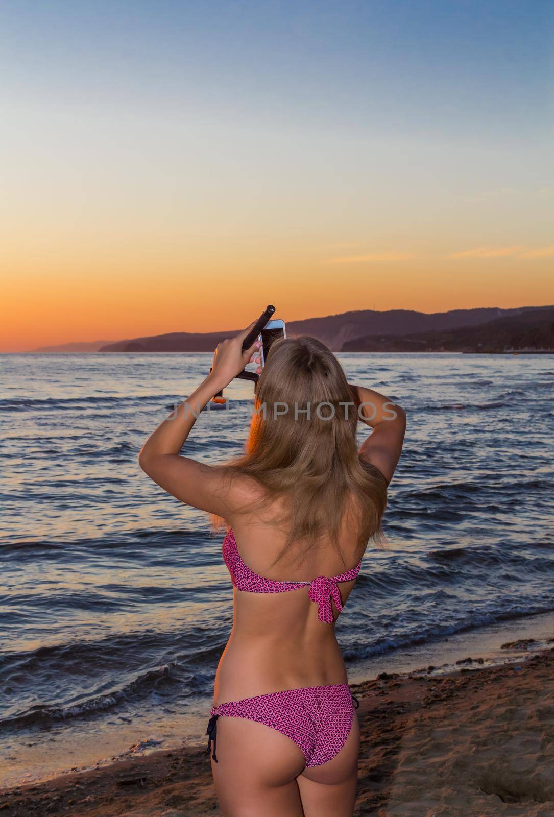 A beautiful young girl in a pink bikini shoots the sunset on the phone camera. Setting sun