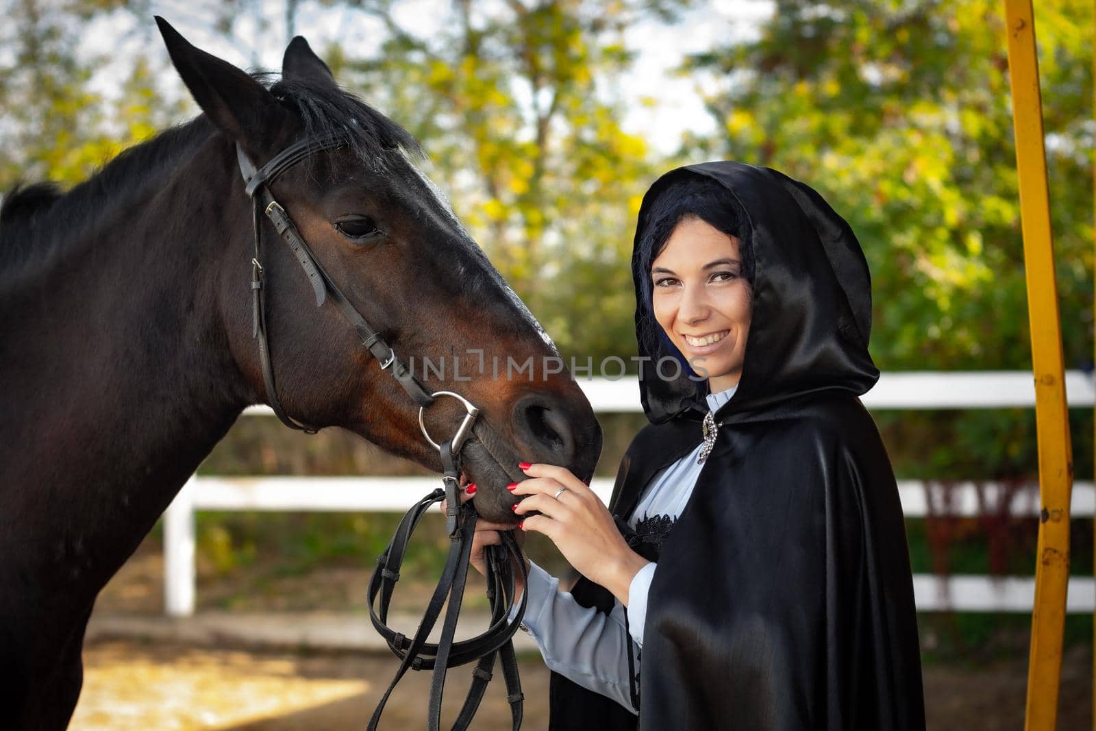 Happy girl in medieval clothes and a black cloak stands with a horse horse on the background of trees and a fence by Madhourse