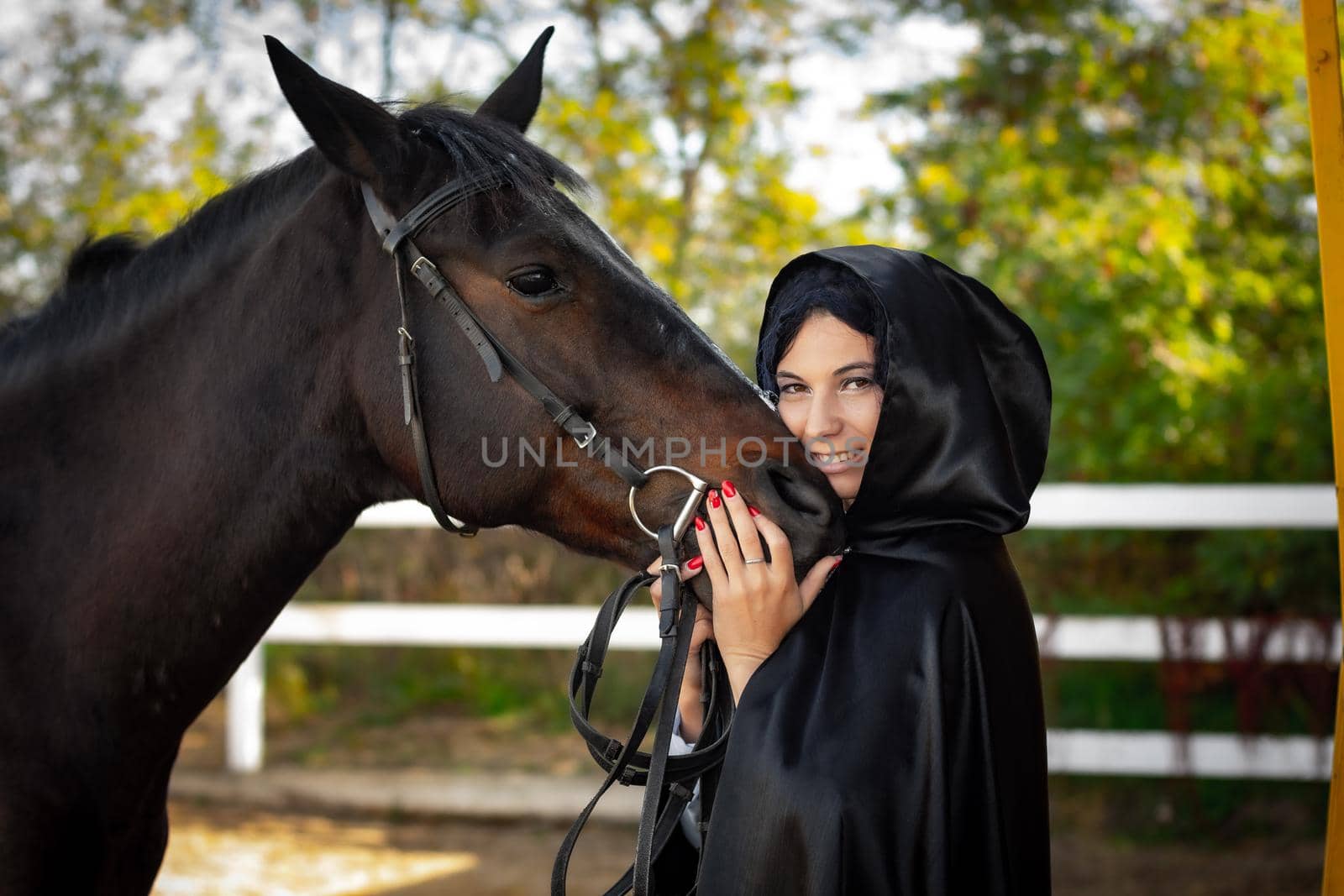 A girl in a black cloak hugs the muzzle of a horse against the background of trees and a fence by Madhourse