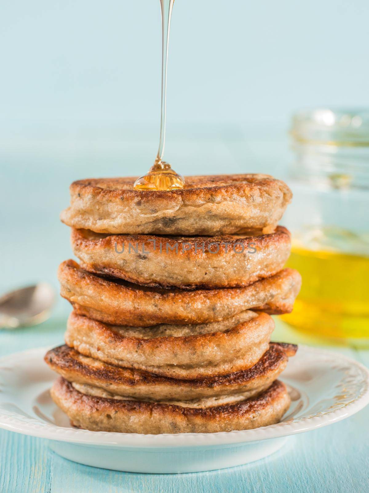 Stack of small pancakes with banana and chia seeds in honey on light blue background. Honey pouring over stack of mini pancakes with chia or poppy seeds.