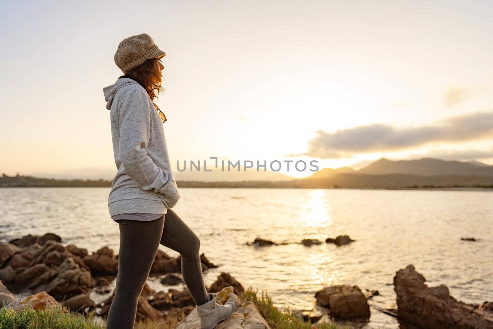 Alone pensive woman standing on sea rocks watching at sunset or sunrise between mountain on the horizon. Casual pretty person with wool hat finds her spirituality living the nature in travel vacation by robbyfontanesi