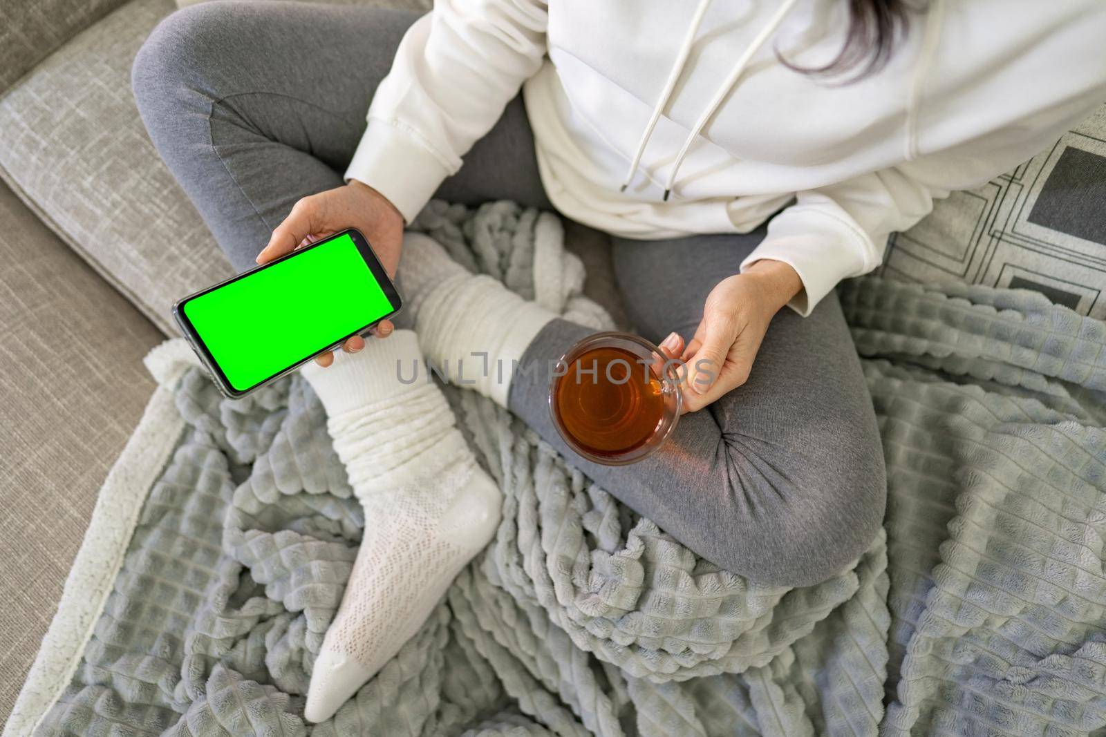 Young person spending time at home using smartphone to chat in sovial network. View from top of bored young woman holding cell with green screen display copy space and tea mug having fun with internet by robbyfontanesi