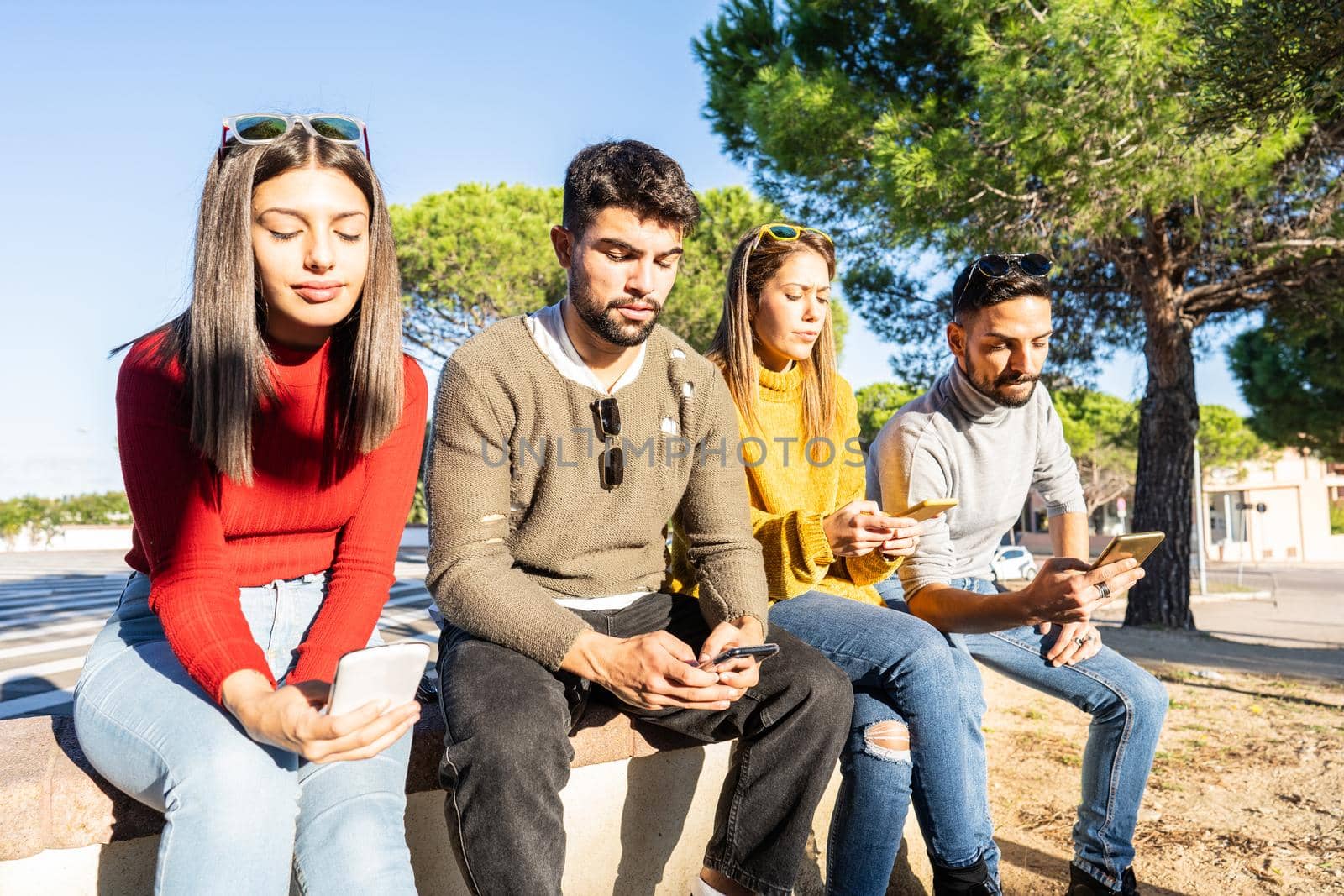 Group of serious young people sitting on a city wall spending time using smartphone with mobile connection. New normal human habits and mutual relations due to internet social network addictions