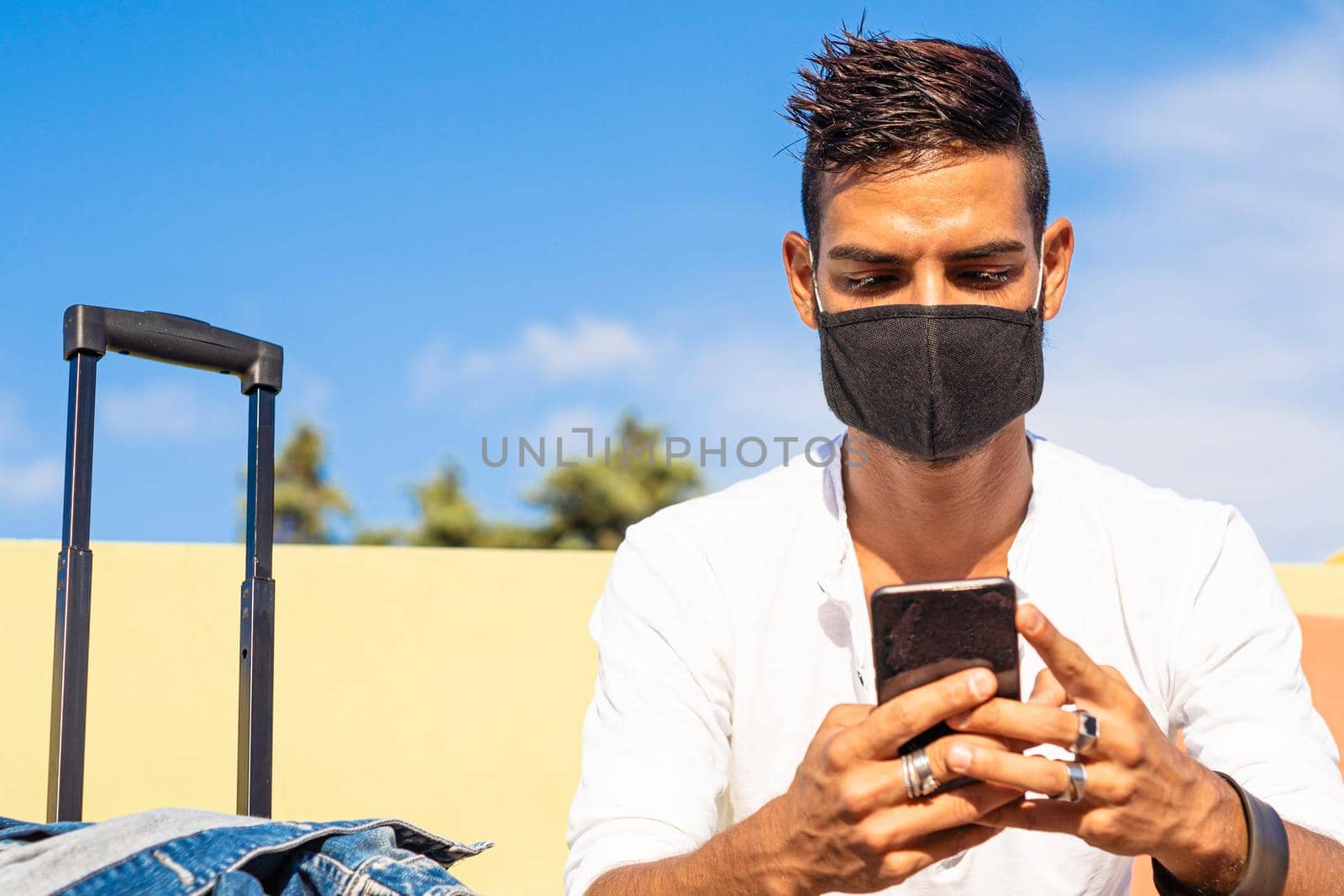 Front close up view of young solo traveller man sitting on a bench wearing protective black mask to cover face against Coronavirus pandemic using smartphone waiting bus with trolley bag next to him