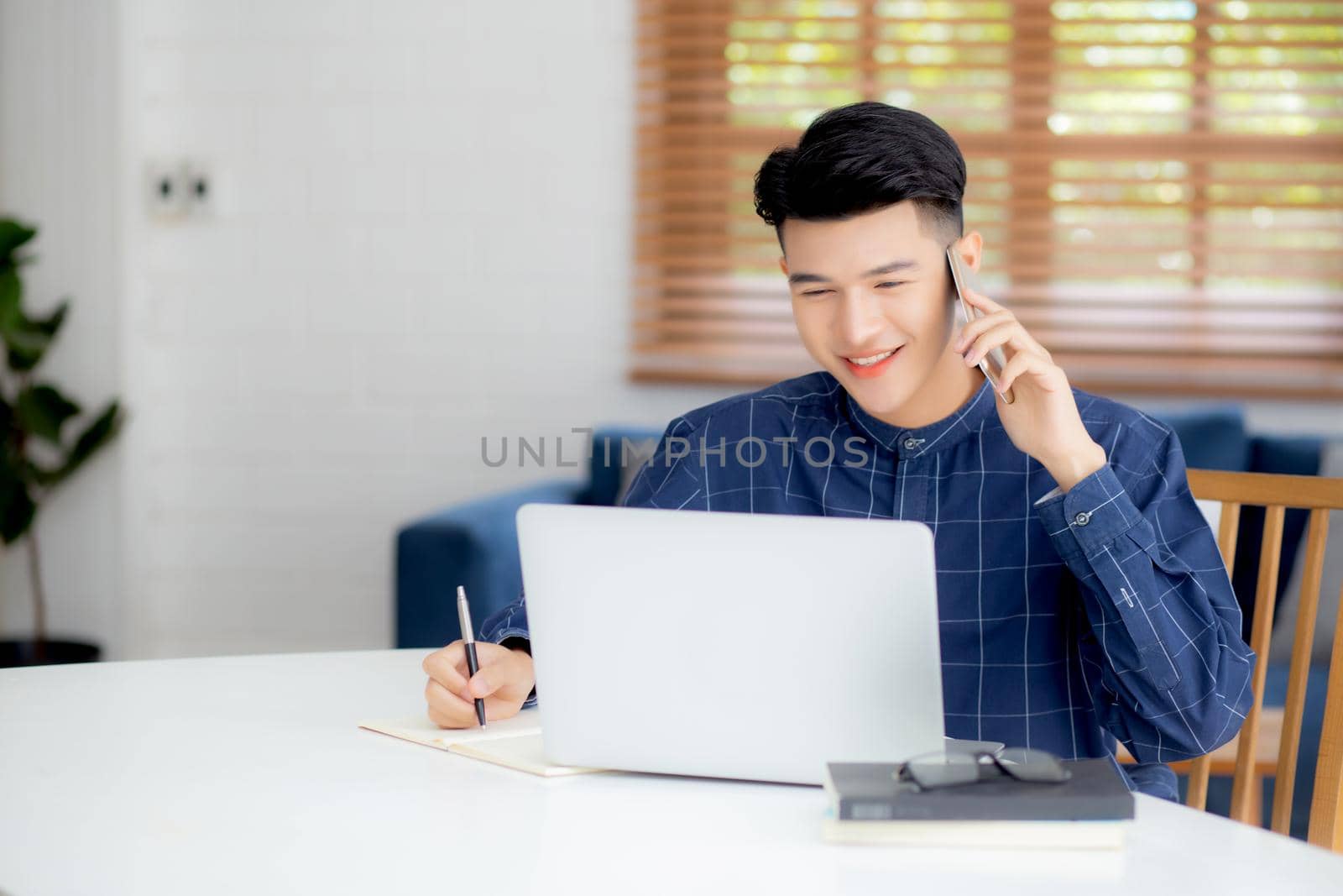 Young asian man talking phone and work from home with laptop computer, freelance male writing on notebook and speak on smartphone, stay home, domestic life, business and communication concept.