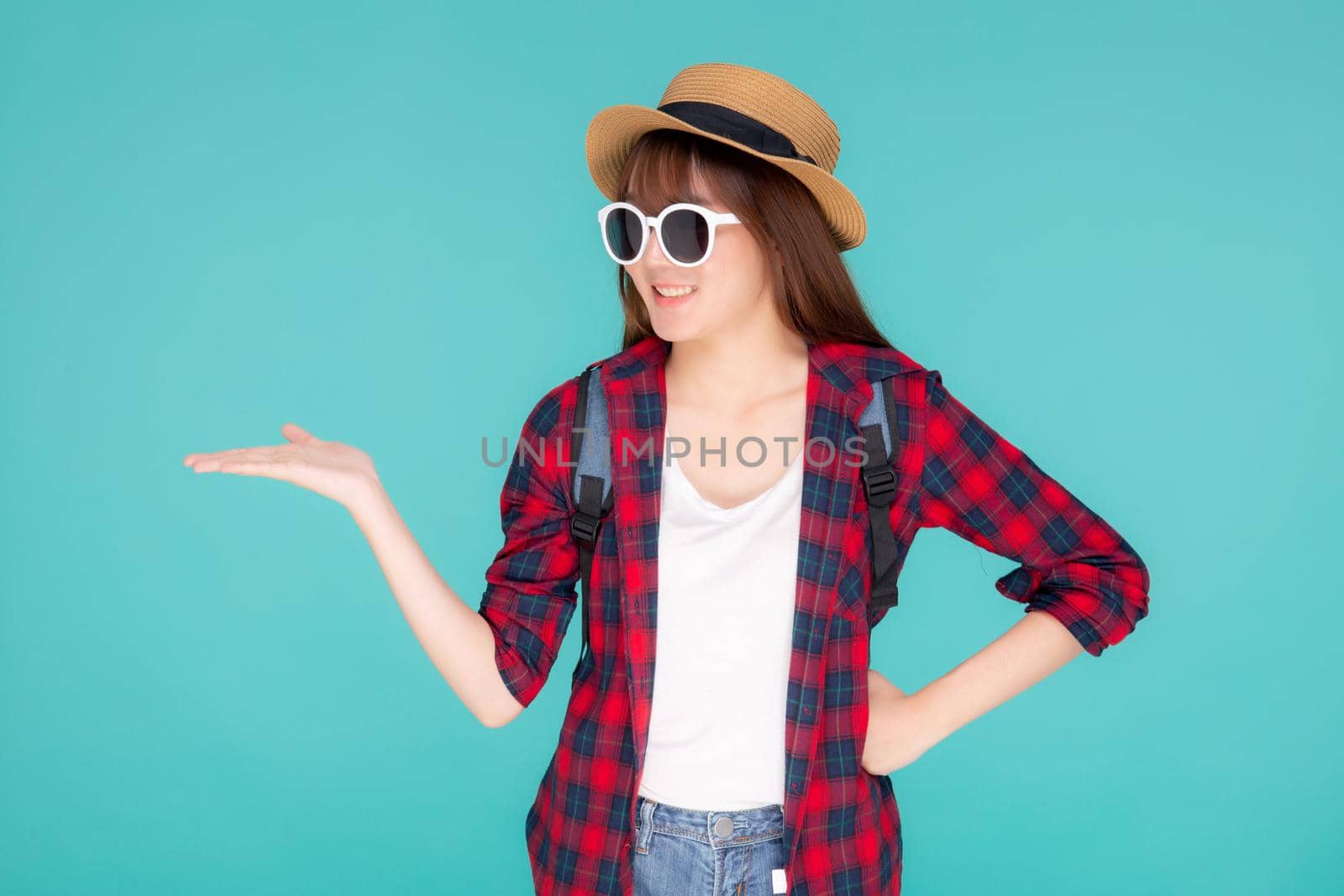 Beautiful portrait young asian woman wear sunglasses and hat smile and enjoy in summer gesture presenting something isolated on blue background, tourist girl in vacation travel concept.