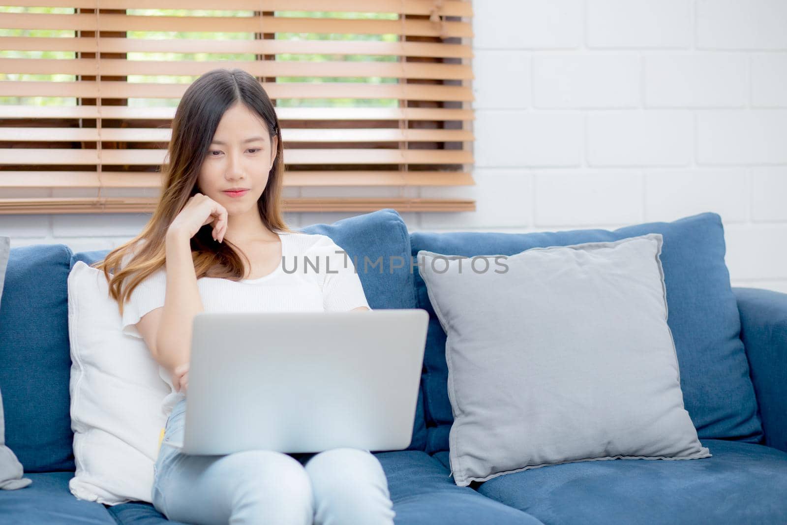 Young asian business woman smile and work from home with laptop computer online to internet on sofa in living room, freelance girl using notebook on couch with comfort, new normal, lifestyle concept.
