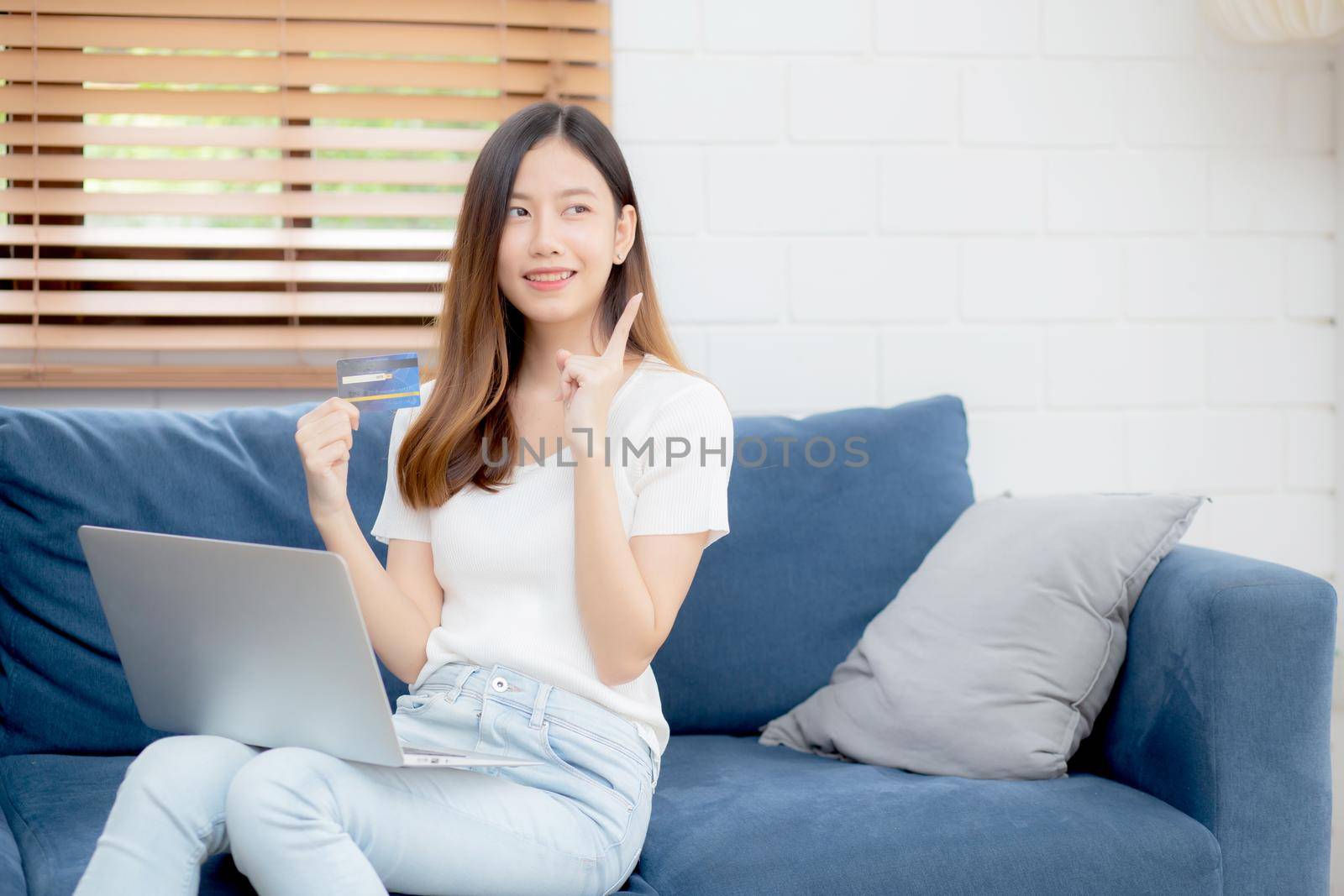 Young asian woman sitting thinking idea using credit card with laptop computer on couch, girl shopping online for buy and payment with notebook on sofa, finance and debit, lifestyle concept.