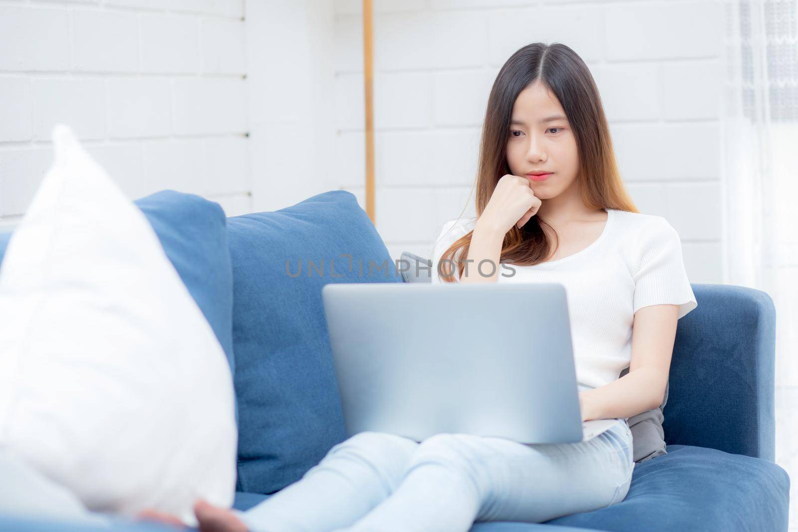 Young asian business woman smile and work from home with laptop computer online to internet on sofa in living room, freelance girl using notebook on couch with comfort, new normal, lifestyle concept.