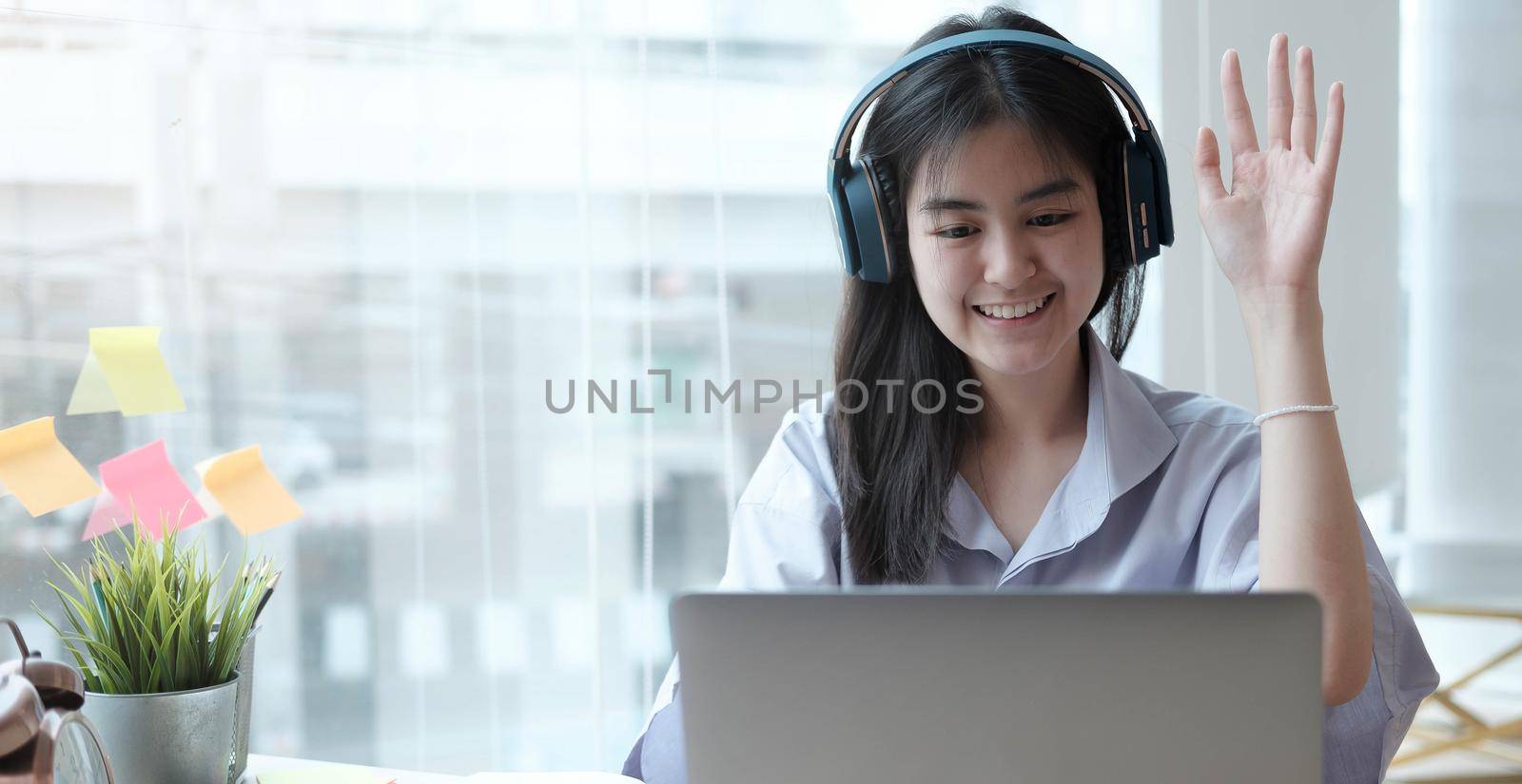 Happy girl school pupil wearing headphones raising hand distance learning online at virtual lesson class with teacher tutor on laptop by zoom video conference call at home. by wichayada