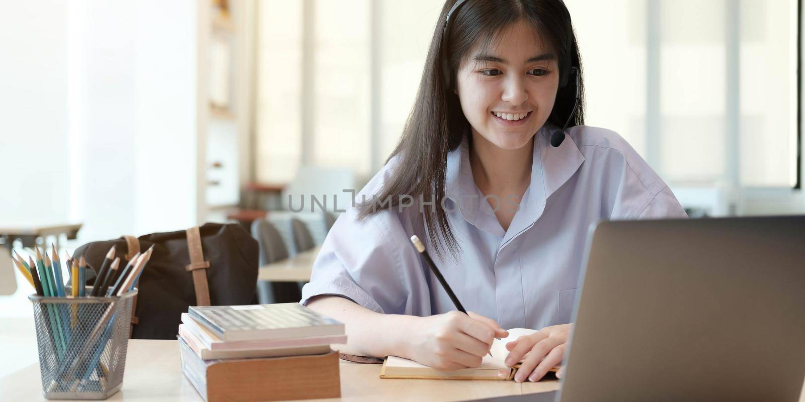 Happy young thai girl with wireless headphones looking at laptop screen, reading listening online courses, studying remotely from home due to pandemic corona virus world outbreak, quarantine time. by wichayada