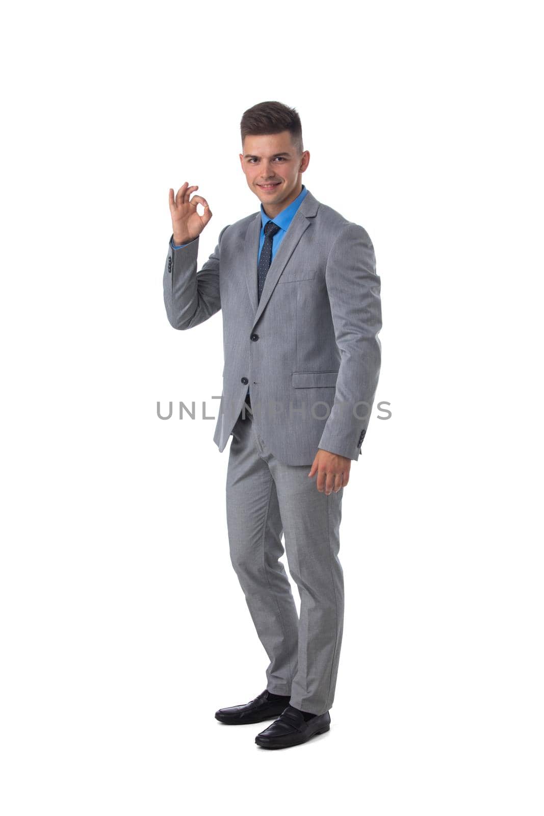 Full length portrait of a young business man in gray suit showing ok sign isolated on white background