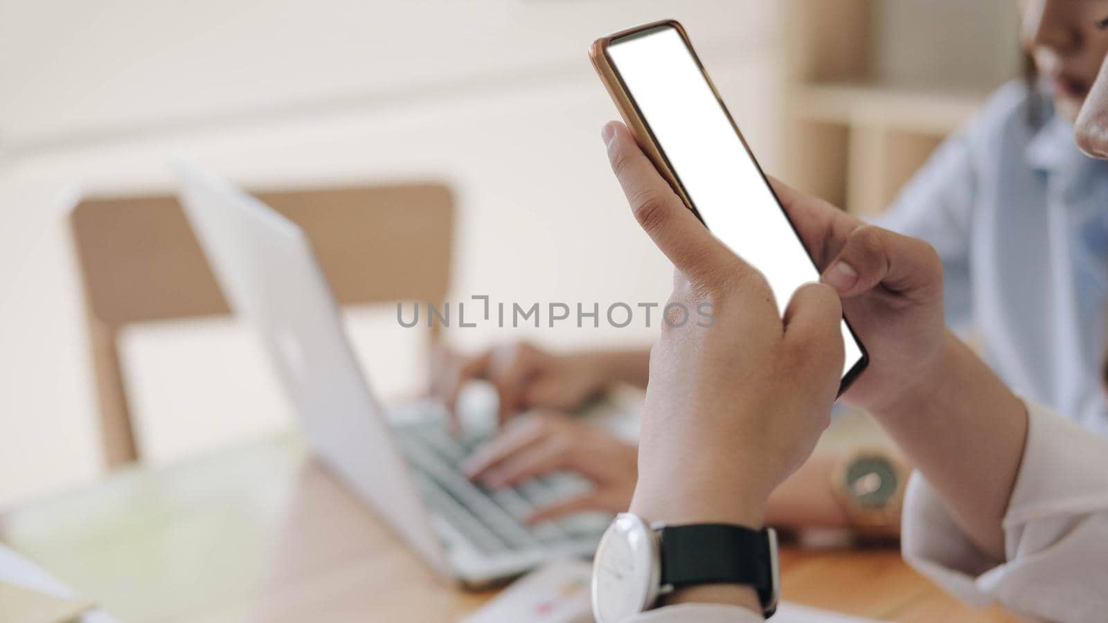 Close-up of smartphone with blank screen in hands of young woman sitting at white table and touching screen. by wichayada