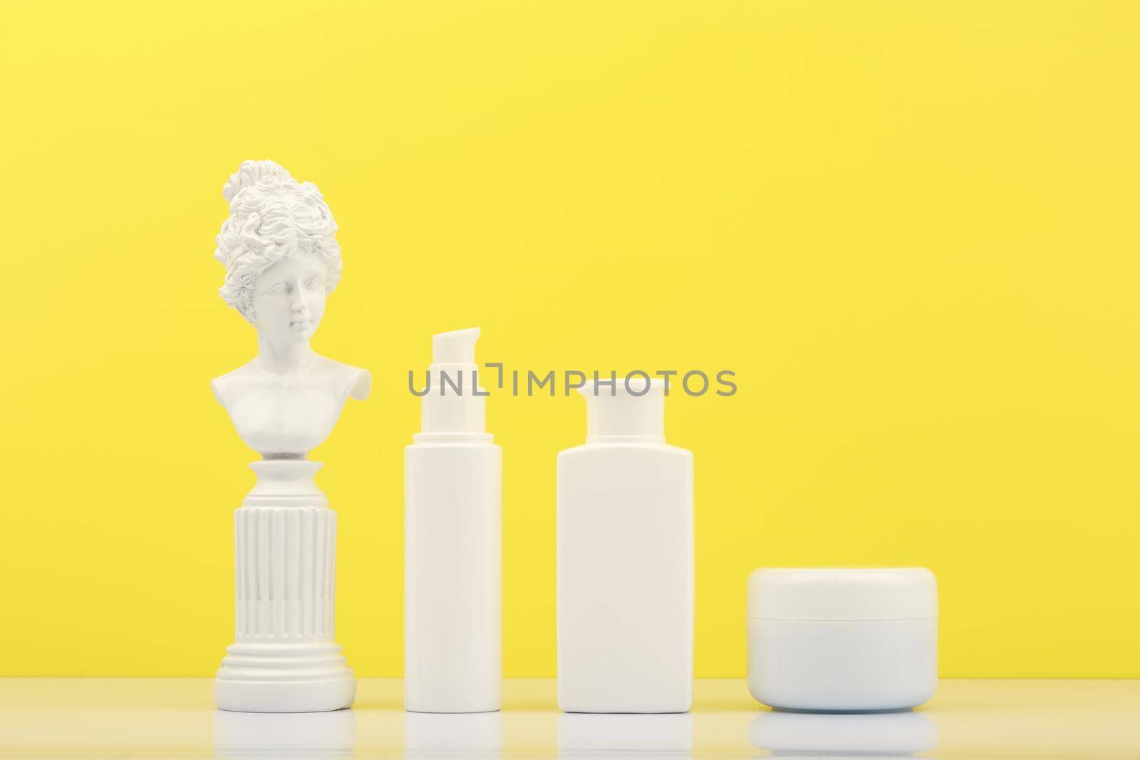 Set of beauty products for daily skin care on white table against bright yellow background with white gypsum statue. Unbranded cosmetic bottles for daily skin routine