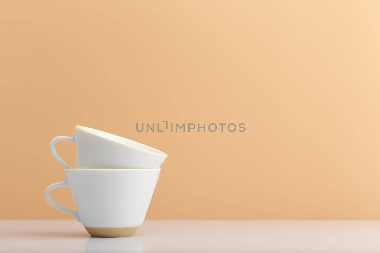 Two white ceramic cups one in another on white table against light beige background with copy space. Concept of hot drinks, tea and coffee