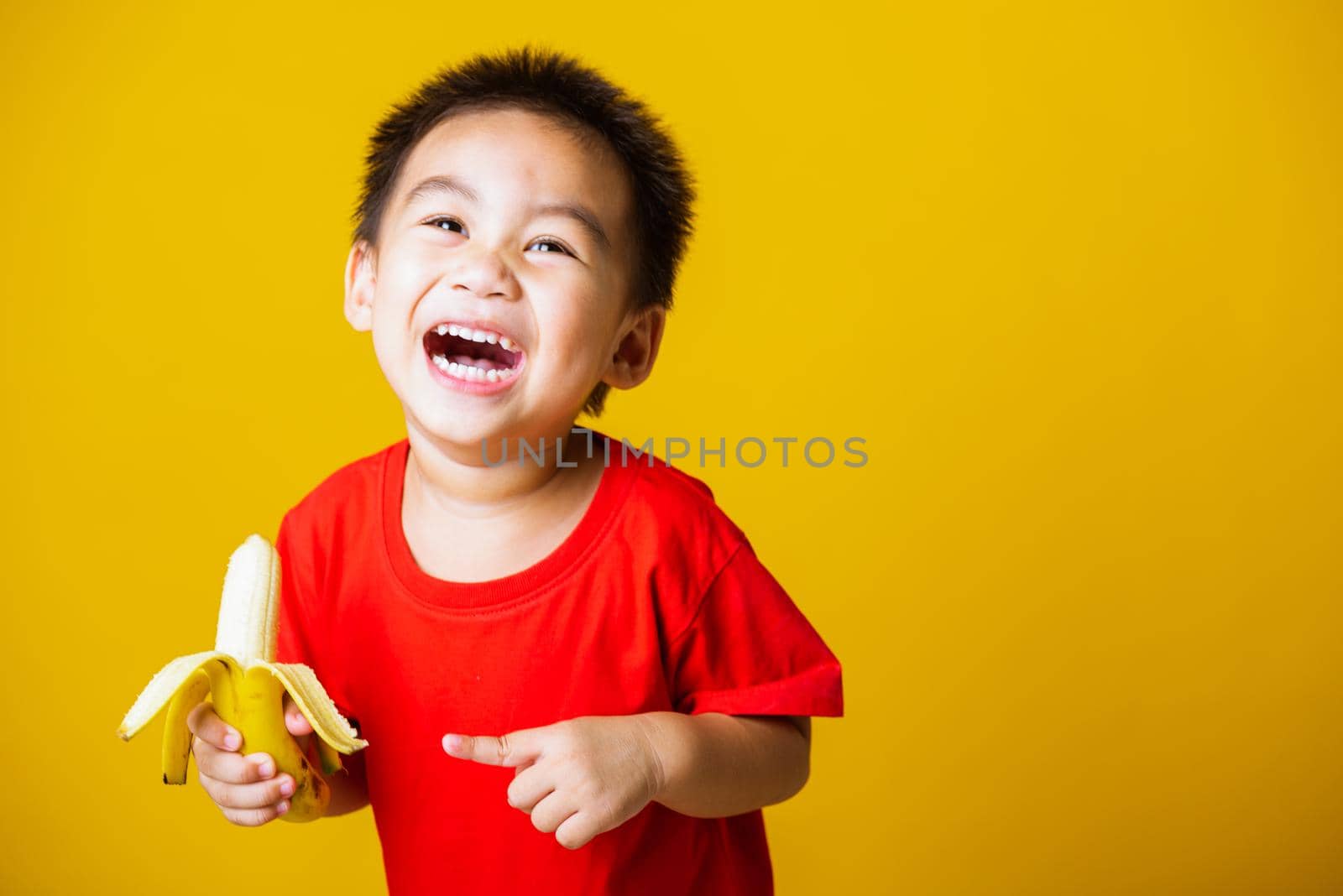 Happy portrait Asian child or kid cute little boy attractive smile wearing red t-shirt playing holds peeled banana for eating, studio shot isolated on yellow background