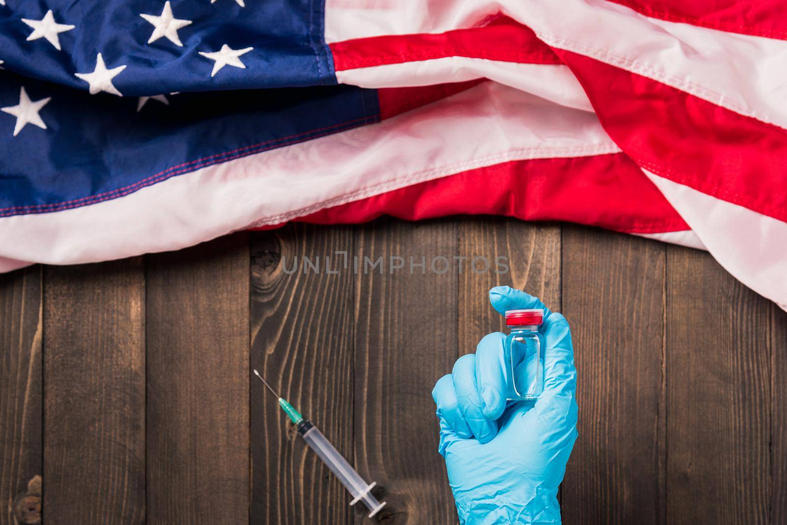 Hands of a doctor wearing gloves holding coronavirus (COVID-19) vial vaccine and syringe with flag United States of America on wood background, USA Vaccination
