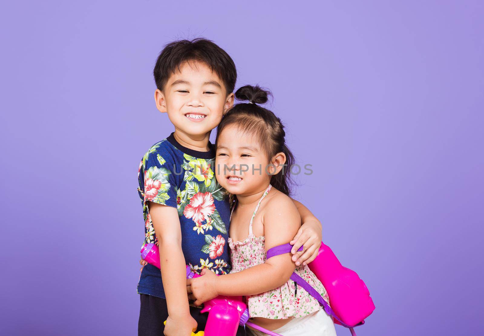 Two Happy Asian little boy and girl holding plastic water gun, Thai children funny hold toy water pistol and smile, studio shot isolated on purple background, Thailand Songkran festival day culture.