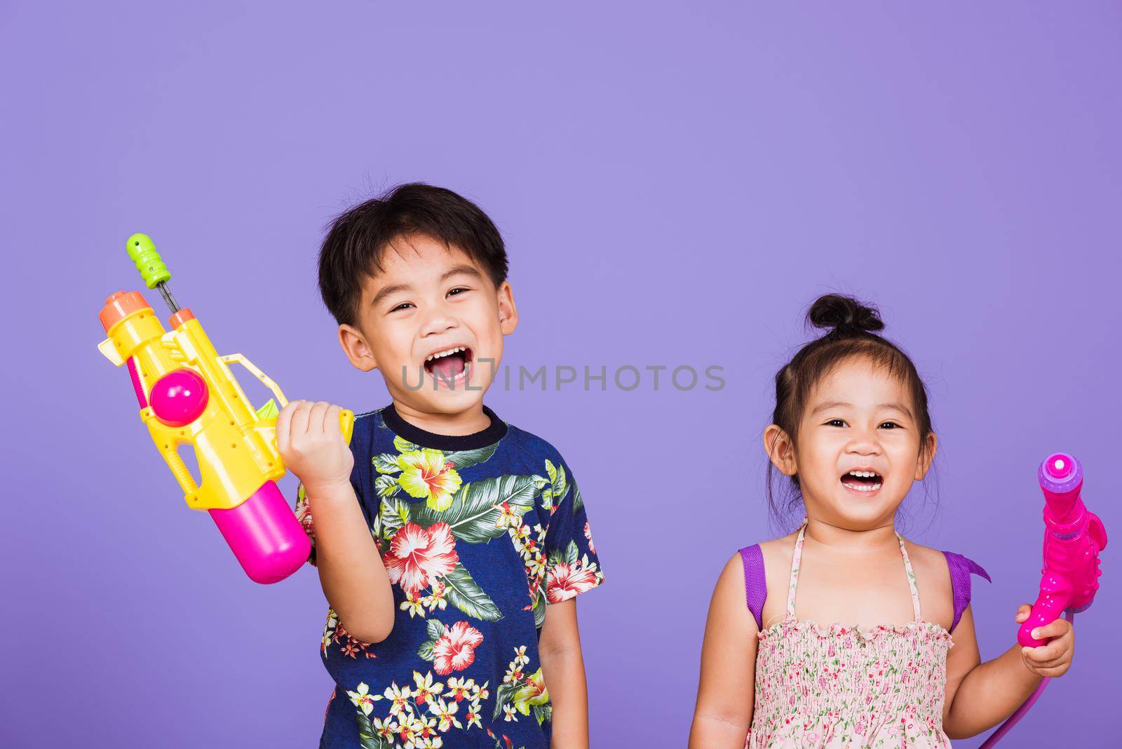 Two Happy Asian little boy and girl holding plastic water gun, Thai children funny hold toy water pistol and smile, studio shot isolated on purple background, Thailand Songkran festival day culture.