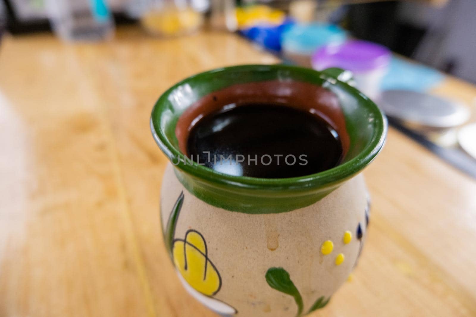 Mexican clay cup filled with coffee on wooden kitchen counter with blurry background. Traditional flowered green and gray craft mug on wood counter. Mexican handcrafts