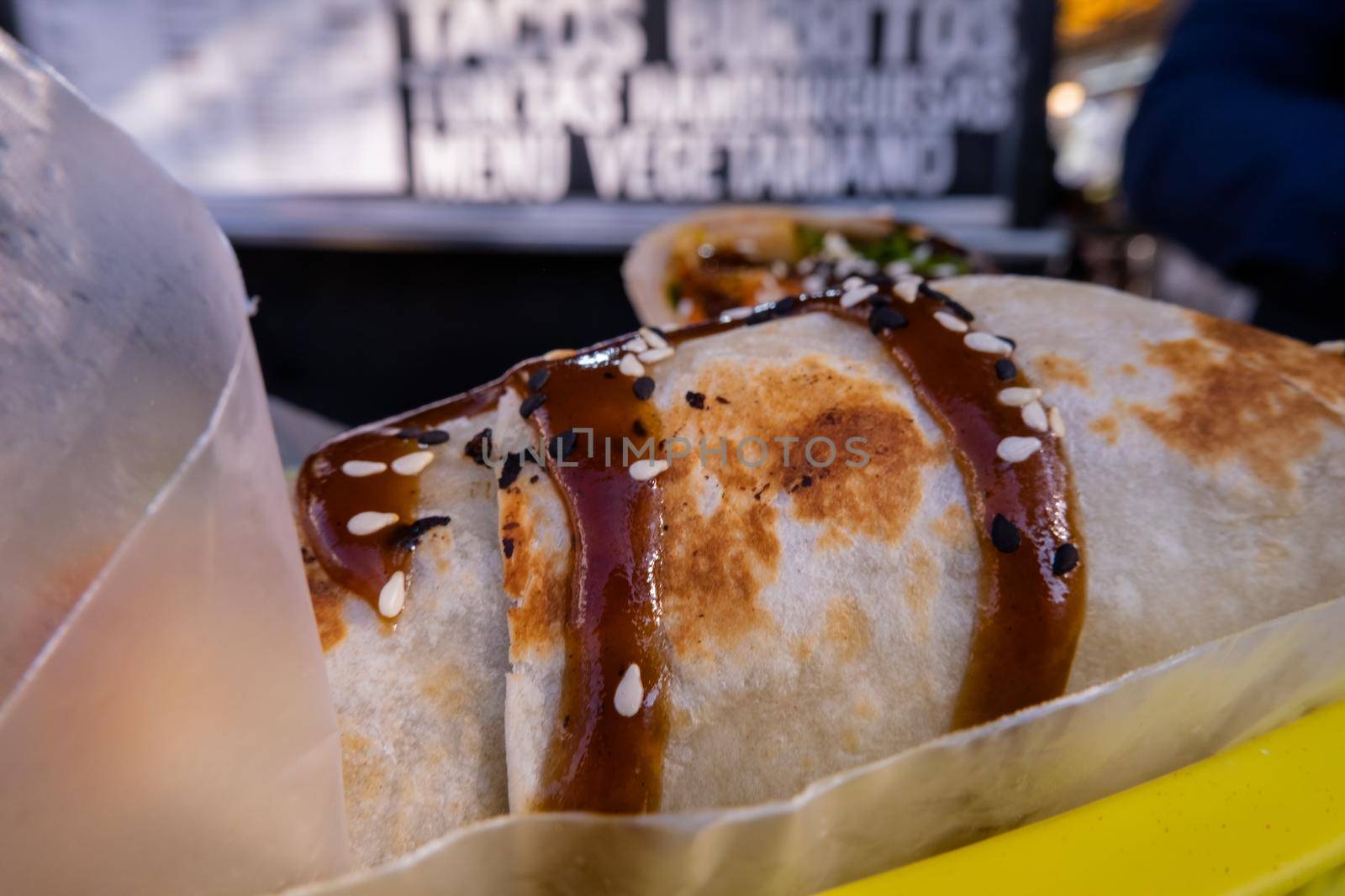 Two vegetarian burritos covered with sauce and seeds with blurry background. Vegan Mexican meal up-close with blurry food truck as background. Traditional Mexican cuisine