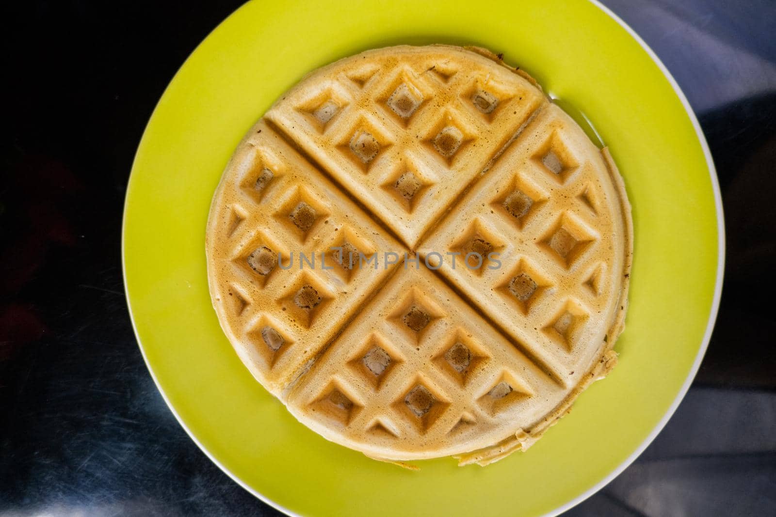 Fresh round waffle on yellow plate from above. Top view of tasty-looking waffle without toppings above black table. Sweet healthy breakfast
