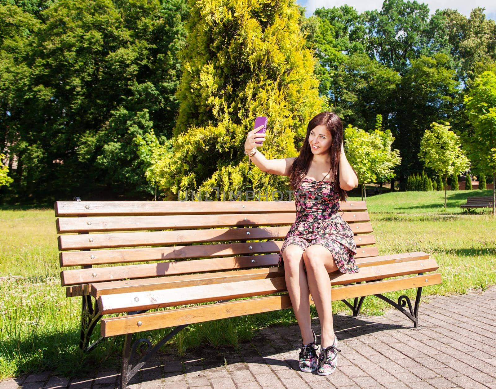 young beautiful brunette makes selfie sitting on a bench in a city park on sunny summer day