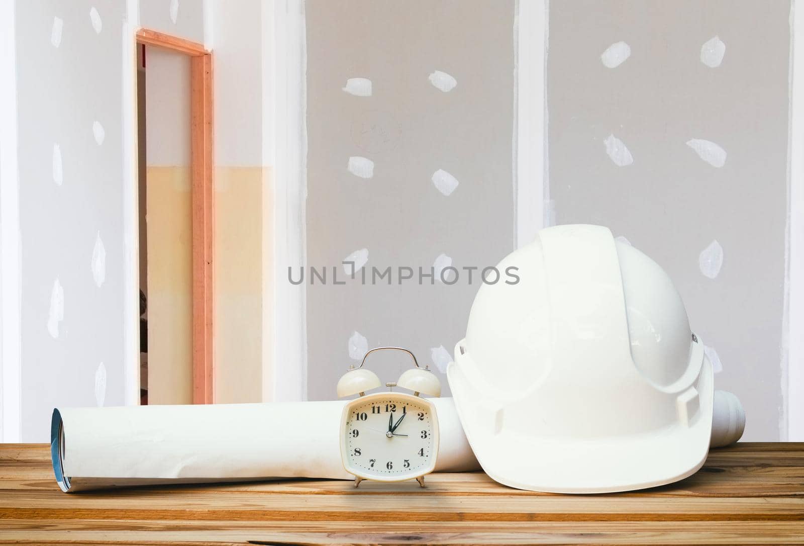 white safety helmet plastic, paper roll plan blueprint alarm clock time a rest at noon on wood floor table and gypsum board wall remodel interior with copy space add text