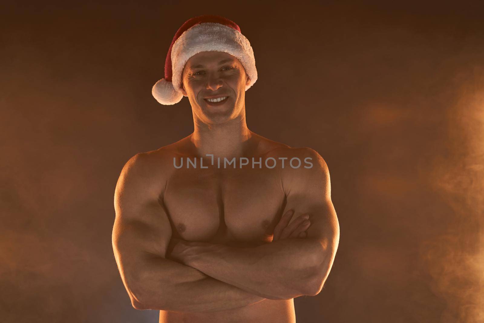 Portrait of muscular man wearing Christmas Santa hat, folded hands and smile on smoky background Macho Shirtless Naked torso stripper Hot sexy Santa guy Seductive male for holiday