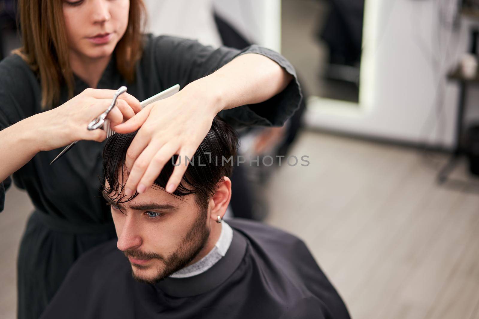 Handsome blue eyed man sitting in barber shop. Hairstylist Hairdresser Woman cutting his hair. Female barber. by andreonegin