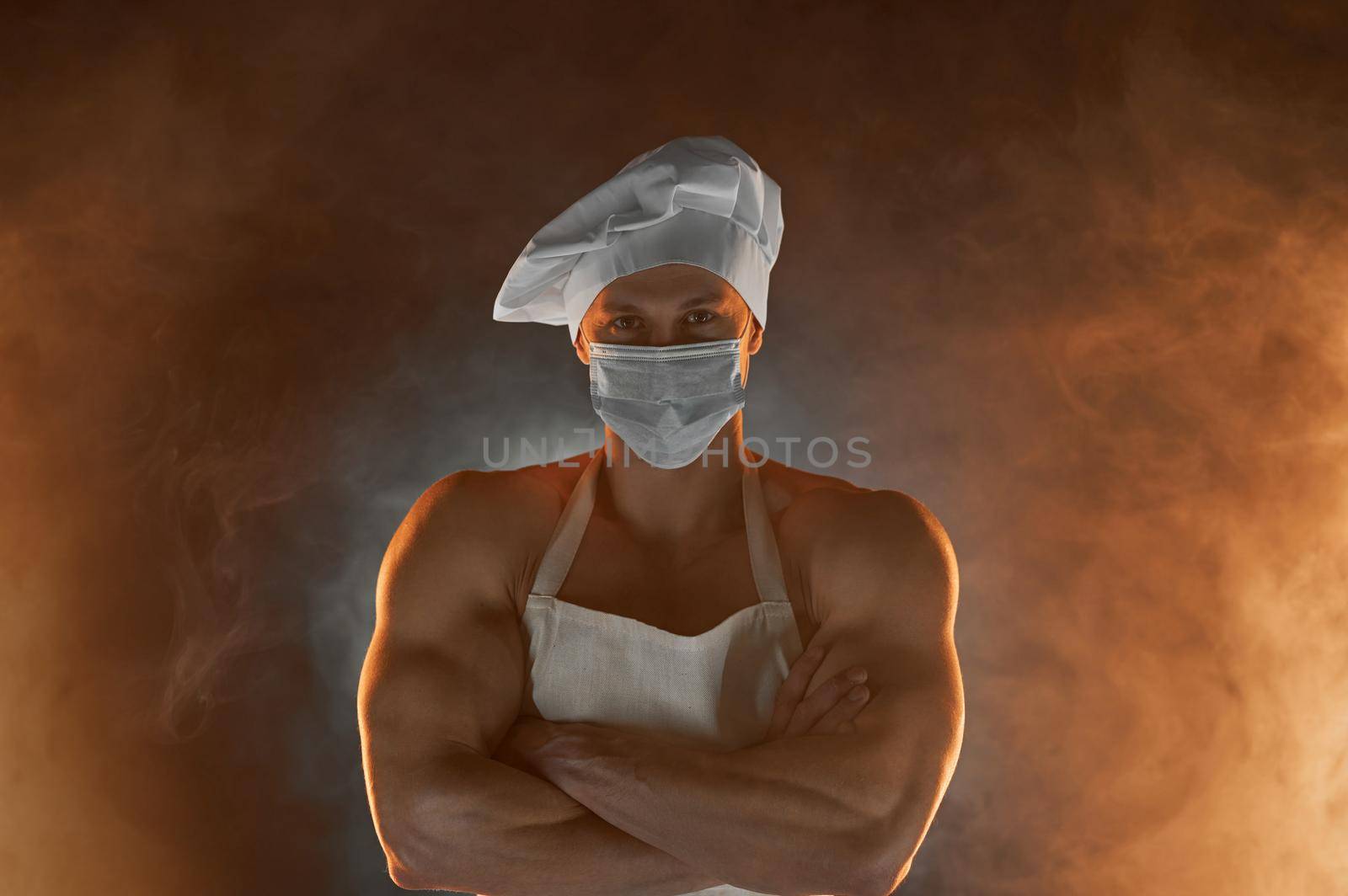 New normal concept. Portrait of muscular chef wearing protective medical mask white apron and chef hat, posing with folded arms on smoky background male housewife. husband in kitchen. brutal butcher.