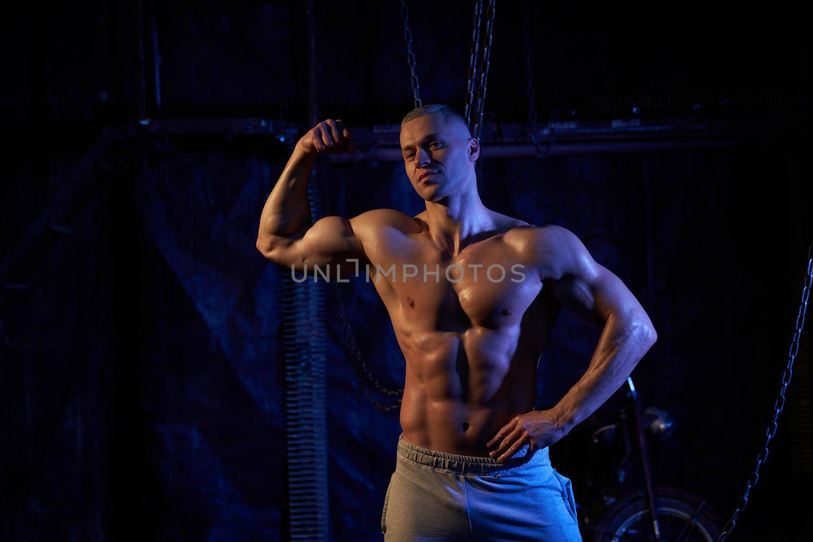 Young shirtless muscular man standing among metal chains, looking seriously at camera, copy space with his back to the camera shows biceps