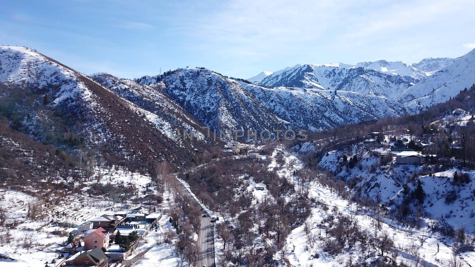 View from the height of the winter road. Along the road there are trees and mountainous terrain. Passing cars, green bus. The shadow of the trees falls on the snow. The house is visible.