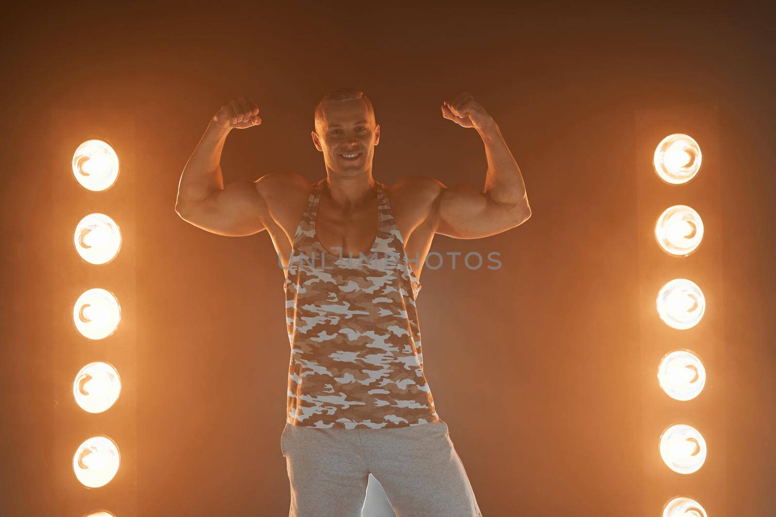 Active lifestyle concept. Professional bodybuilder showing biceps and smiling at camera, lamps illumination and smoke on background