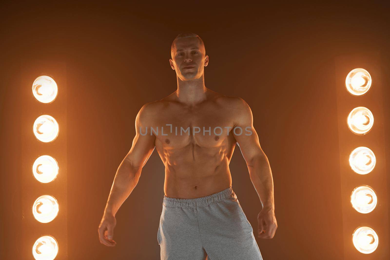 Active lifestyle concept. Professional bodybuilder showing perfect muscular body smiling at camera, lamps illumination smoke on background