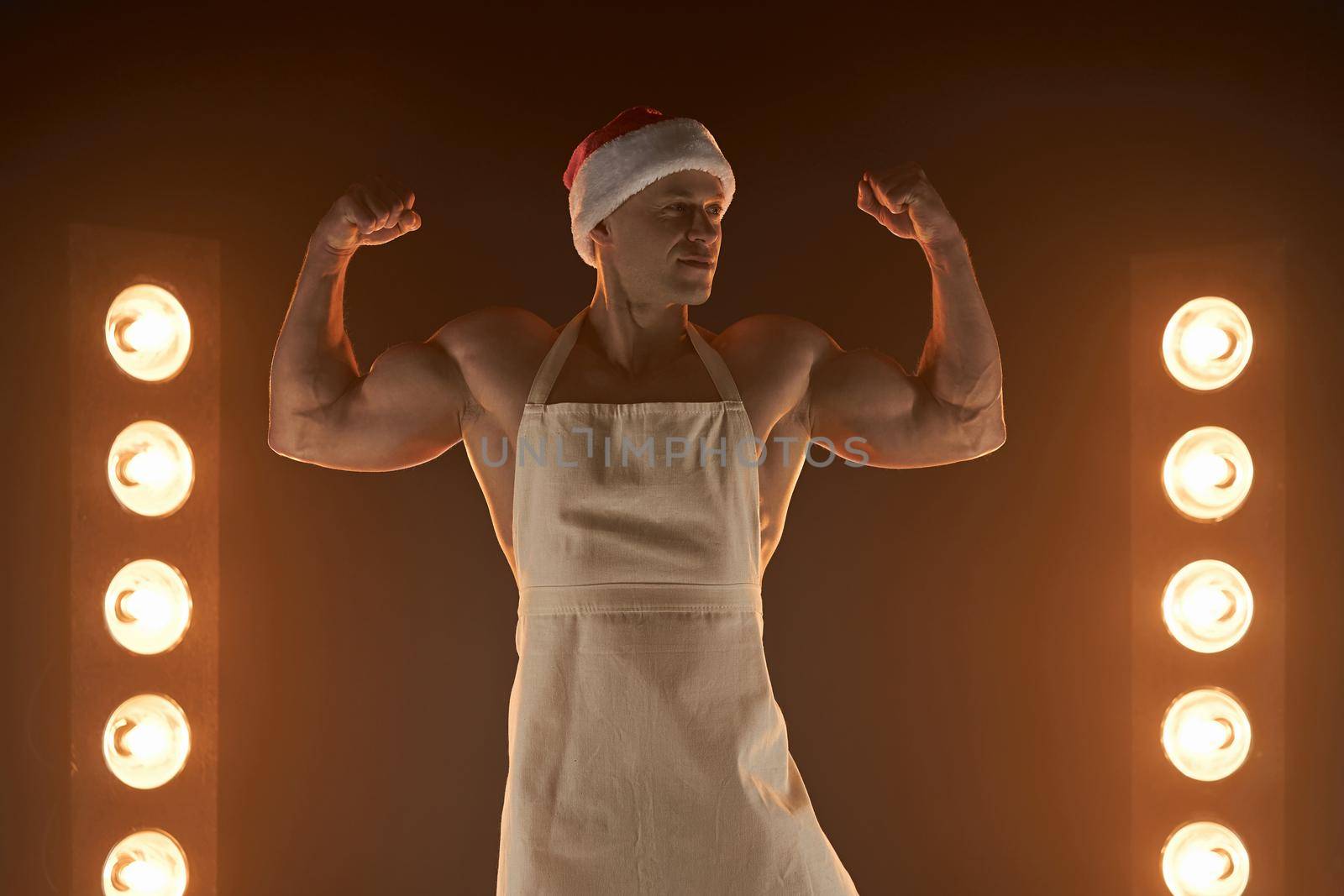 Hot Christmas dessert Sexy muscular Chef wearing apron and Santa hat show biceps smiling, lamps illumination and smoke on background cuisine. male housewife. husband in kitchen. brutal butcher.