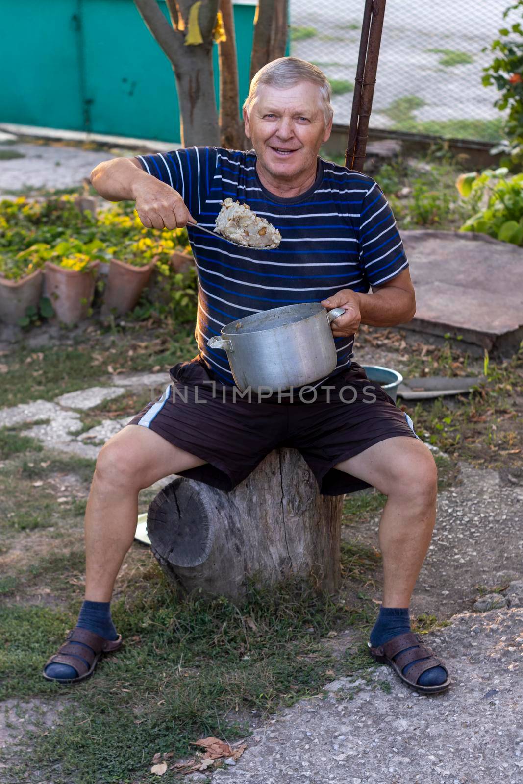 Old man with blond hair eats porridge from pot. Sunny autumn day. Front view. by Essffes
