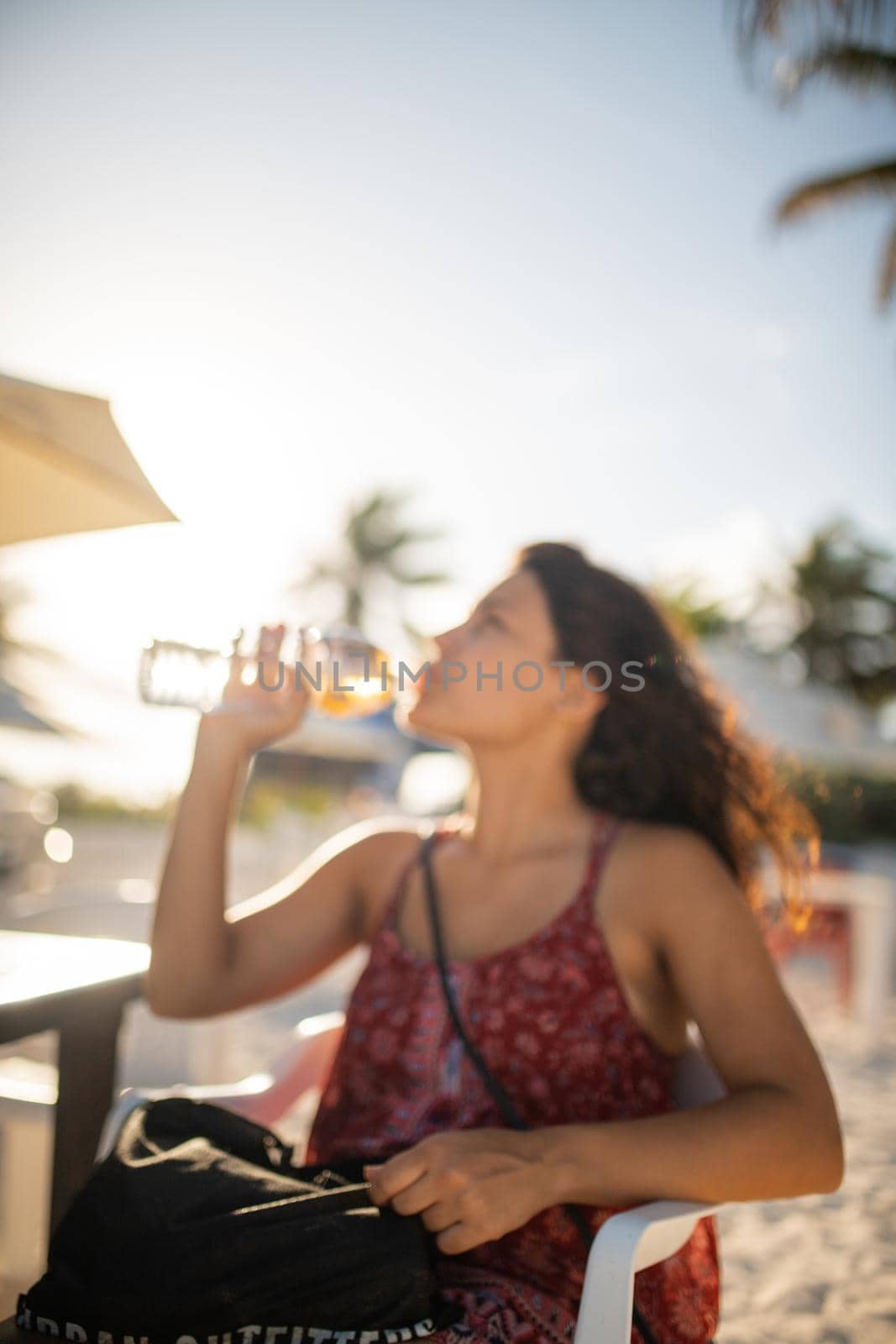 Brunette woman on beach sitting on chair and drinking water from bottle. Portrait of woman drinking water with bright blue sky as background.Tropical summer vacations