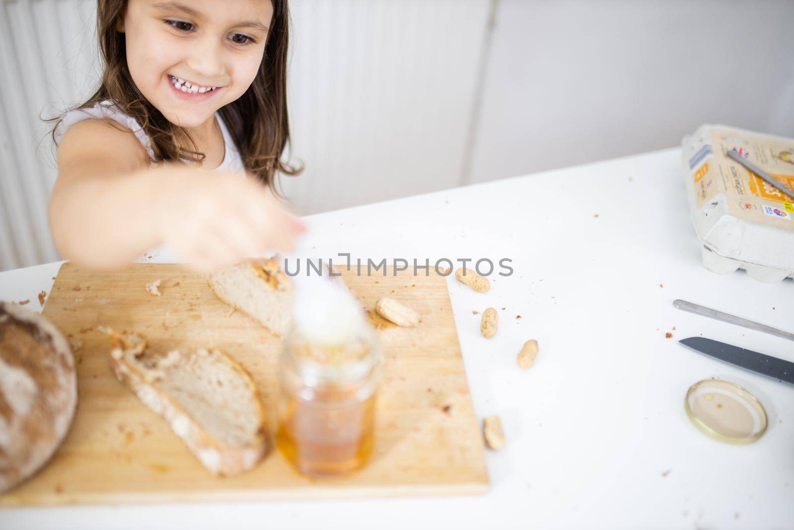 Happy little girl picking honey from jar next to a slice of bread above wooden cutting board. Young child at white table sticking a spoon into honey jar. Sweet and light breakfast
