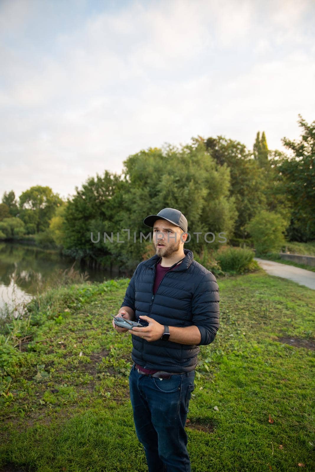 Concentrated man standing next to lake and holding remote control. Man in park with trees and boats on lake as background. People interacting with technology outside