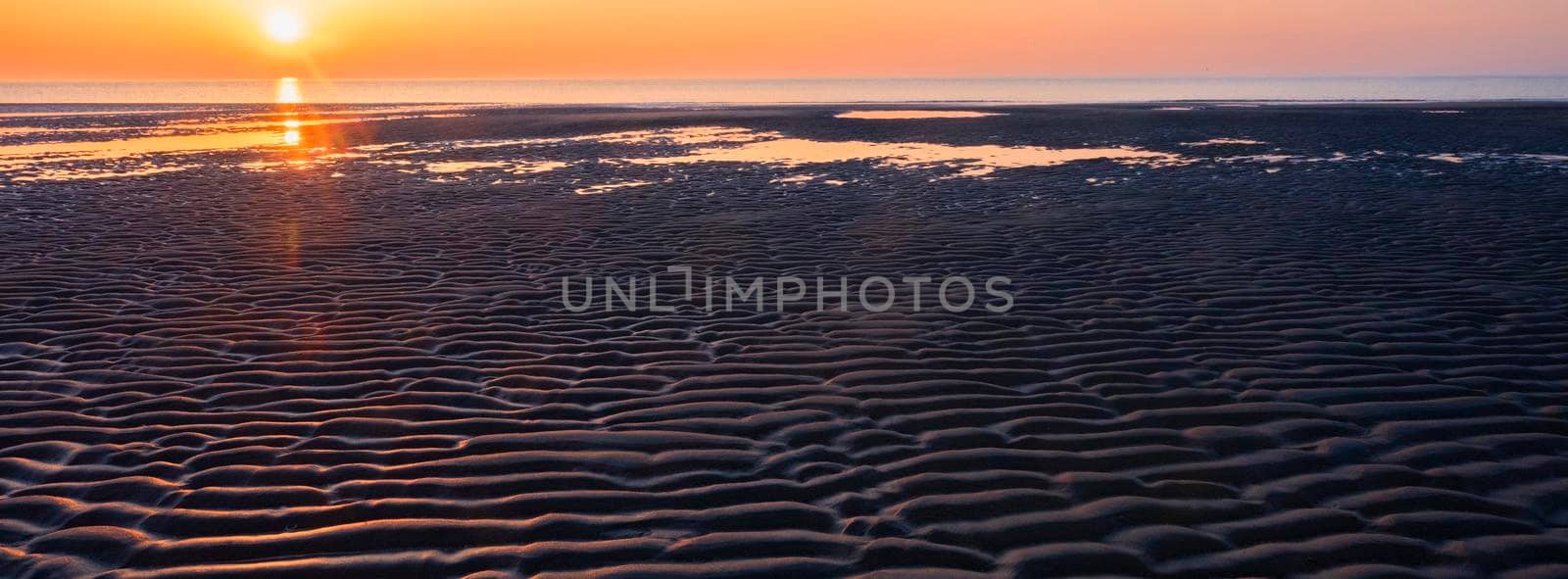 pattern in sand and colorful reflection of setting sun in water by ahavelaar