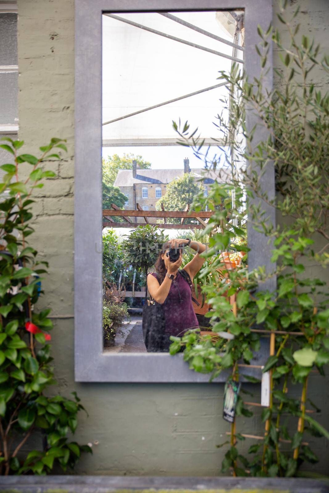 Young brunette woman taking picture of herself in window surrounded by plants. Reflection of woman taking selfie with house and plants as background. Self-portraits and nature