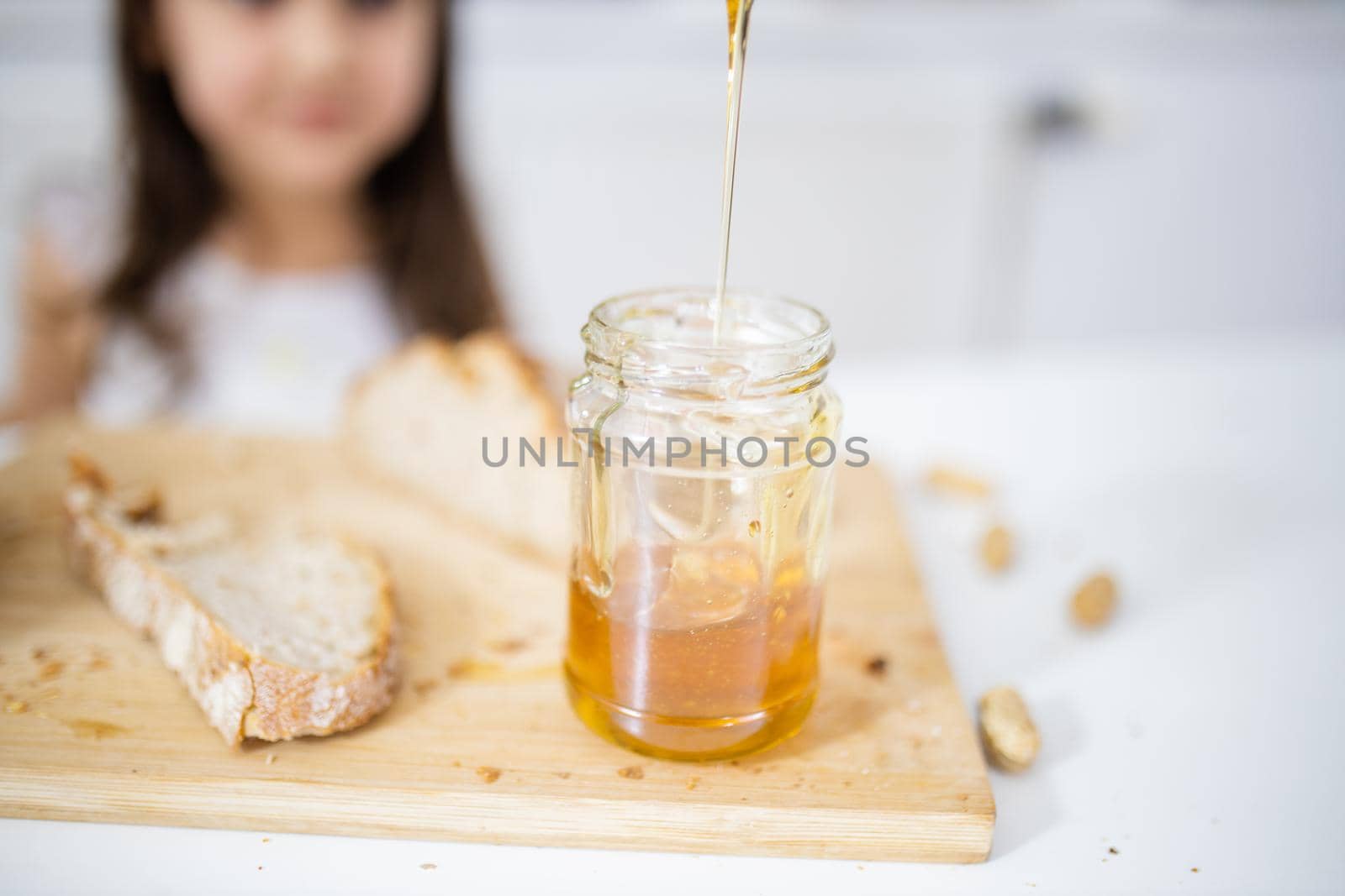 Picking honey from jar next to a slice of bread above wooden cutting board. Young girl at white table watching a honey jar.Sweet and light breakfast