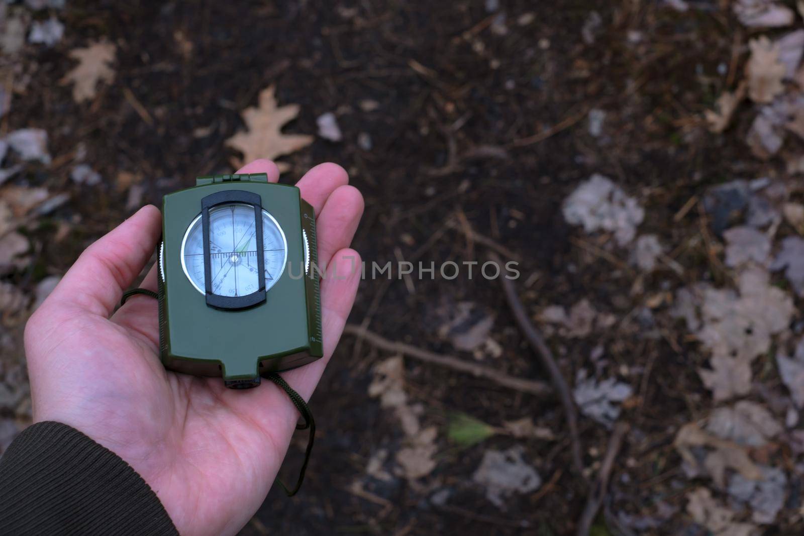 A hand holds a green tactical prismatic compass over a forest trail. The cover is closed.