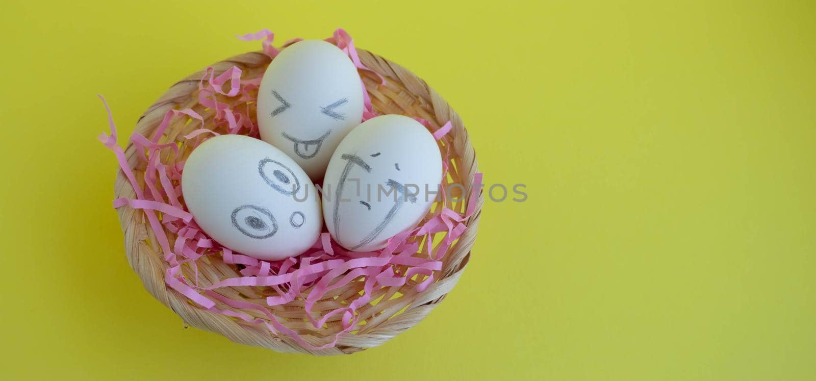 On the pink tinsel in the basket are three charming white eggs with faces on a yellow background. Easter Concept. Place for your text.
